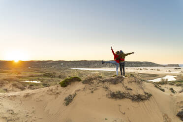 Young carefree couple with hand raised standing by man on sand dune during sunset - SBOF02704