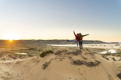 Junges sorgloses Paar mit erhobener Hand neben einem Mann auf einer Sanddüne bei Sonnenuntergang - SBOF02704