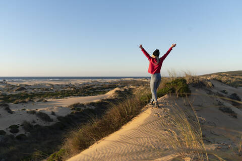 Sorglose Frau mit erhobener Hand, die bei Sonnenuntergang auf einer Sanddüne spazieren geht, lizenzfreies Stockfoto