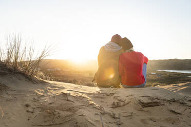 Woman leaning on man shoulder while sitting on sand dune during sunset - SBOF02709