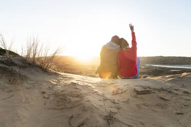 Woman with hand raised showing peace sign while sitting with man on sand dune - SBOF02710