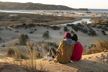 Paar mit Strickmütze sitzt bei Sonnenuntergang auf einer Sanddüne zusammen - SBOF02711