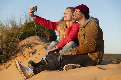Lächelndes Paar, das ein Selfie mit dem Handy macht, während es auf einer Sanddüne sitzt - SBOF02712