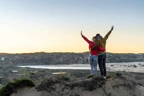 Paar steht mit Arm um und Hand erhoben auf Sanddüne während Sonnenuntergang, lizenzfreies Stockfoto