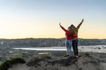 Couple standing with arm around and hand raised on sand dune during sunset - SBOF02724