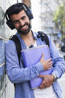 Happy businessman with documents listening music while leaning on wall - PNAF00753