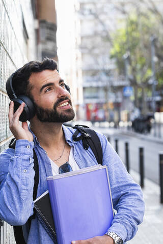 Männlicher Unternehmer mit Dokumenten, der in die Richtung schaut, während er Musik über drahtlose Technologie hört, lizenzfreies Stockfoto