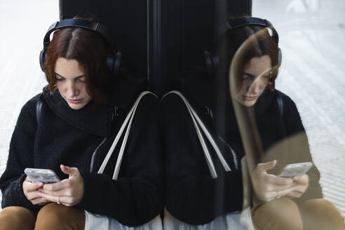 Young businesswoman listening music through wireless headphones while using smart phone by glass wall - PNAF00756