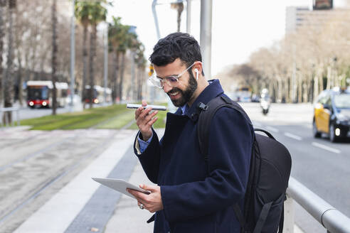 Smiling businessman with digital tablet talking on smart phone through speaker at station - PNAF00763