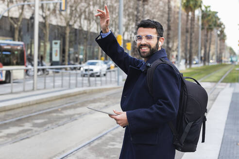 Smiling young businessman with digital tablet waving while standing at station - PNAF00765