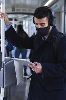 Businessman wearing protective face mask using digital tablet while standing in train - PNAF00766