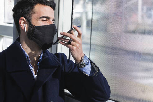 Male business professional wearing protective face mask talking on smart phone through speaker in train - PNAF00770