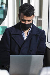 Young businessman wearing protective face mask using laptop while sitting in train - PNAF00771