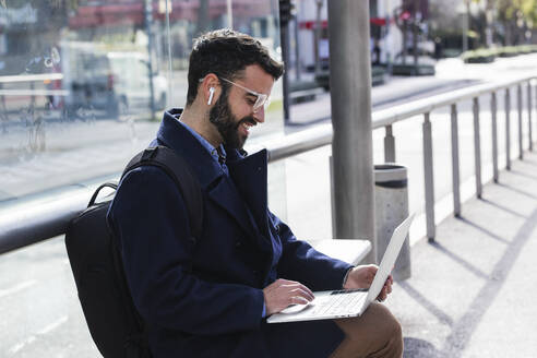 Male freelancer working on laptop while sitting on bench at station - PNAF00773