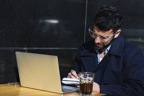 Businessman with laptop writing while sitting at sidewalk cafe - PNAF00774