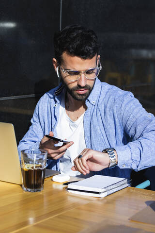 Männlicher Unternehmer mit Mobiltelefon, der in einem Straßencafé sitzt und die Zeit überprüft, lizenzfreies Stockfoto