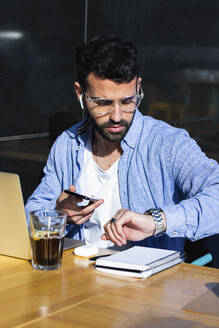 Male entrepreneur with mobile phone checking time while sitting at sidewalk cafe - PNAF00775