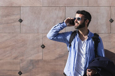 Young businessman talking on smart phone while standing against wall during sunny day - PNAF00777