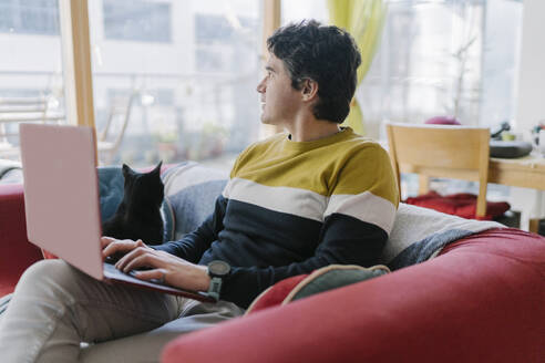 Mittlerer erwachsener Mann, der einen Laptop benutzt und wegschaut, während er zu Hause auf dem Sofa sitzt - BOYF01921