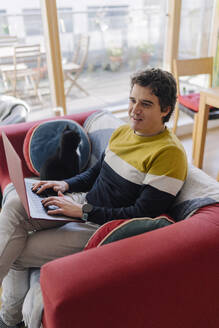 Mann sitzt mit Katze auf Sofa und benutzt Laptop im Wohnzimmer - BOYF01922