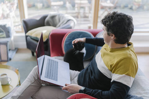 Mid adult man with laptop stroking cat while sitting on sofa at home stock photo