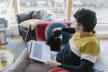 Mittlerer erwachsener Mann mit Laptop, der seine Katze streichelt, während er zu Hause auf dem Sofa sitzt - BOYF01923