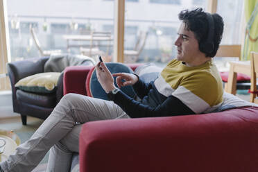 Mann benutzt sein Smartphone, während er zu Hause über drahtlose Kopfhörer Musik hört - BOYF01924