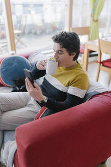 Mittlerer erwachsener Mann, der ein Mobiltelefon benutzt, während er zu Hause auf dem Sofa sitzt und Kaffee trinkt - BOYF01926