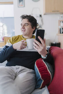 Mann mit Smartphone und Kaffeetasse auf dem Sofa sitzend und wegschauend - BOYF01927