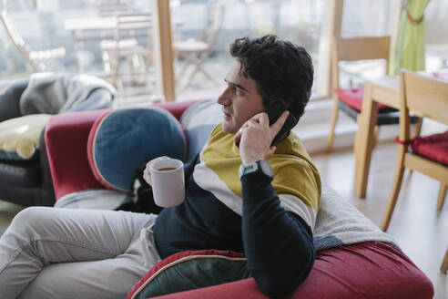 Mittlerer erwachsener Mann mit Kaffeetasse auf dem Sofa sitzend, während er zu Hause mit dem Handy telefoniert - BOYF01928