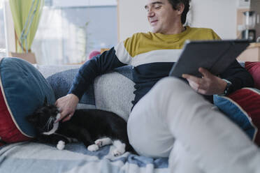 Lächelnder Mann mit digitalem Tablet, der seine Katze streichelt, während er auf dem Sofa im Wohnzimmer sitzt - BOYF01932