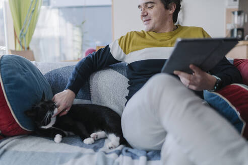 Smiling man with digital tablet stroking cat while sitting on sofa in living room - BOYF01932