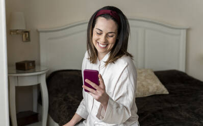 Fröhliche junge Frau, die auf dem Bett sitzend ein Smartphone benutzt - AFVF08228