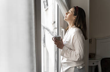 Entspannte junge Frau mit geschlossenen Augen, die eine Kaffeetasse hält, während sie am Fenster steht - AFVF08230