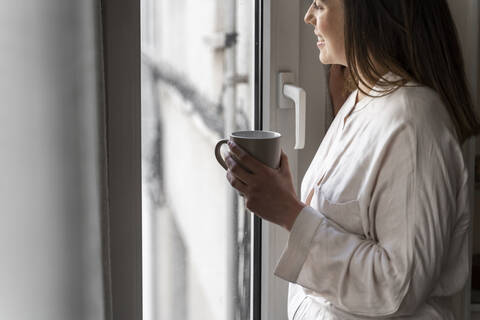 Glückliche junge Frau mit Kaffeetasse, die durch das Fenster ihres Hauses schaut, lizenzfreies Stockfoto