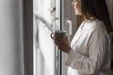 Glückliche junge Frau mit Kaffeetasse, die durch das Fenster ihres Hauses schaut - AFVF08231