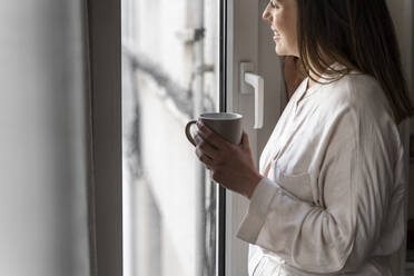 Happy young woman with coffee cup looking through window at home - AFVF08231