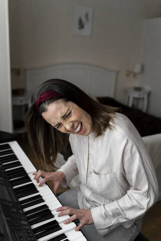 Fröhliche junge Frau singt beim Klavierspielen im Schlafzimmer, lizenzfreies Stockfoto