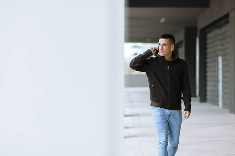 Young businessman talking on phone looking away while walking on footpath stock photo