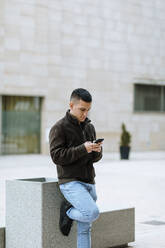 Junger Mann benutzt ein Smartphone, während er an einem Gebäude steht - OCAF00630