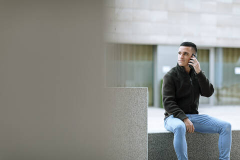 Junger Mann spricht am Telefon, während er auf einer Bank vor einem Gebäude sitzt, lizenzfreies Stockfoto