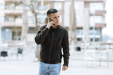 Businessman talking on smart phone while walking against building - OCAF00634