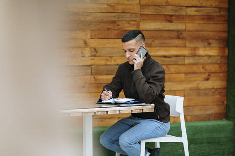 Young male entrepreneur talking on the phone while writing in diary at sidewalk cafe stock photo