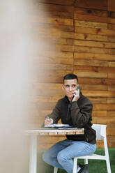 Lächelnder Geschäftsmann, der mit seinem Smartphone spricht und wegschaut, während er in einem Straßencafé sitzt - OCAF00638