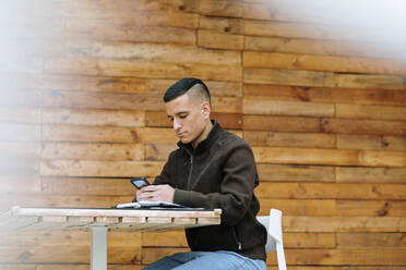 Junger männlicher Unternehmer, der in einem Straßencafé sitzt und sein Smartphone benutzt - OCAF00640
