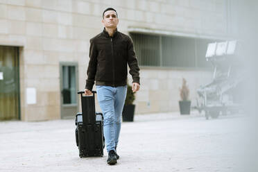 Young man with suitcase walking on the road against building - OCAF00645