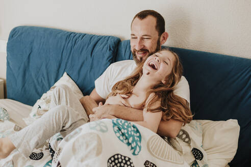 Daughter laughing with father while sitting on bed - GMLF00997