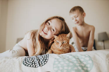 Lächelndes Mädchen, das sich an eine Katze lehnt, während der Bruder im Hintergrund im Schlafzimmer zu Hause ist - GMLF00999