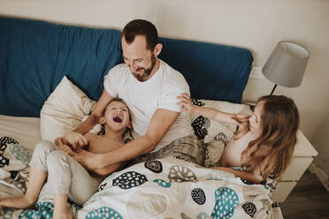 Father playing with cheerful son while daughter sitting on bed at home - GMLF01003