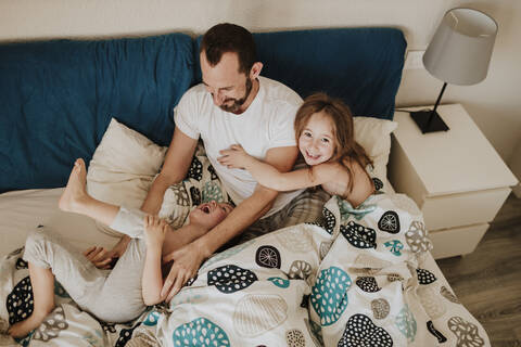 Happy father playing with cheerful son while daughter sitting on bed at home stock photo